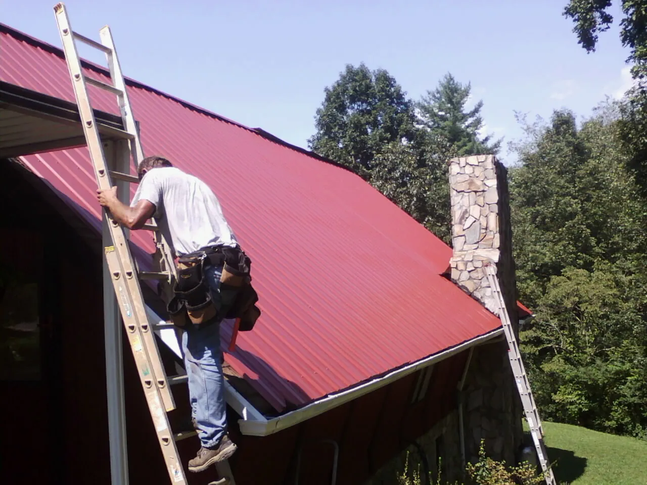 Roofer on a ladder beside a red metal roof and stone chimney — Soffit & Fascia Repair by Roofing Brothers in Boydton