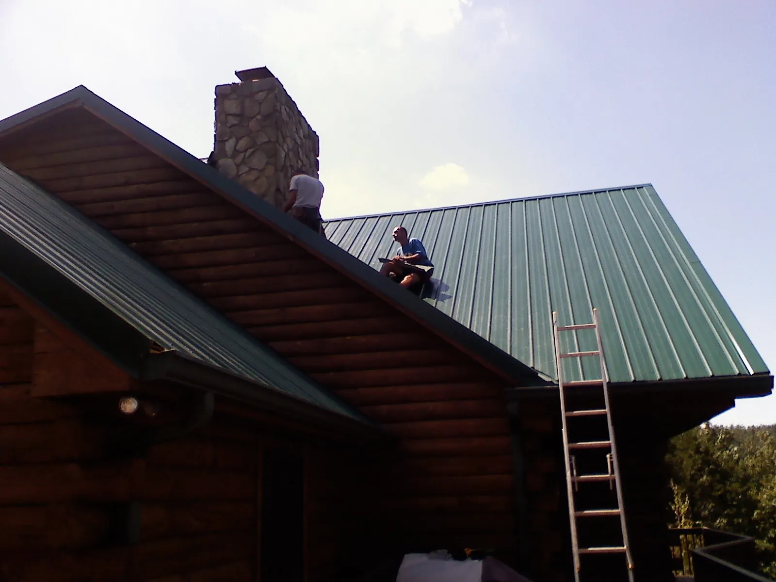 Roofers installing green metal panels beside a stone chimney on a log home in Boydton